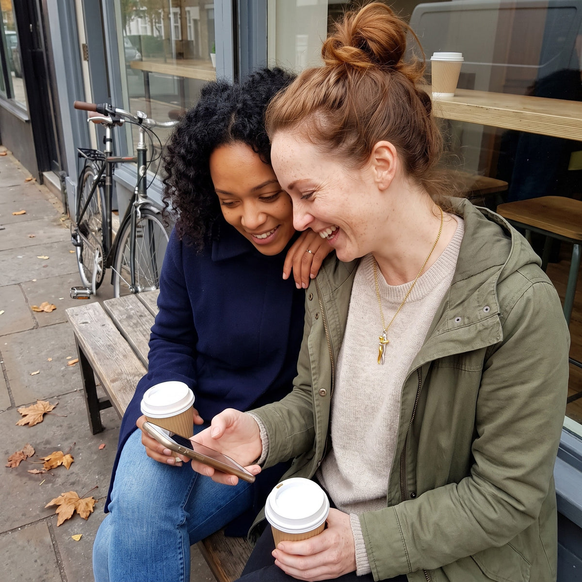 Two friends laughing outside a café, one wearing the Hug Necklace